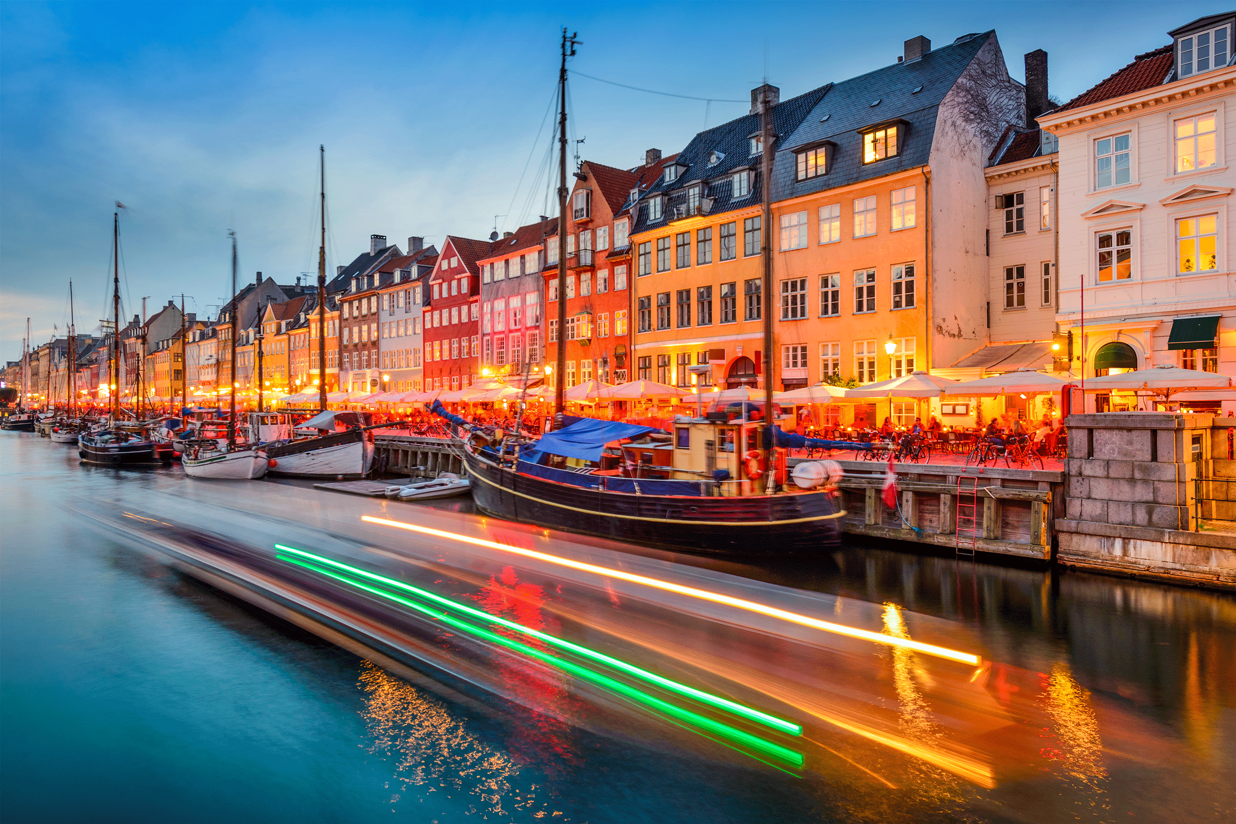 Nyhavn canal in Copenhagen at dusk, with colourful townhouses, wooden boats and light trails on the water.