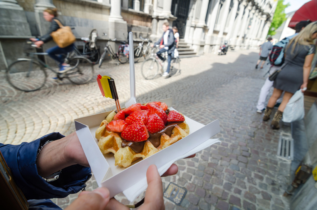 Belgian waffle topped with strawberries and chocolate spread, held in a cobbled street with cyclists passing behind.