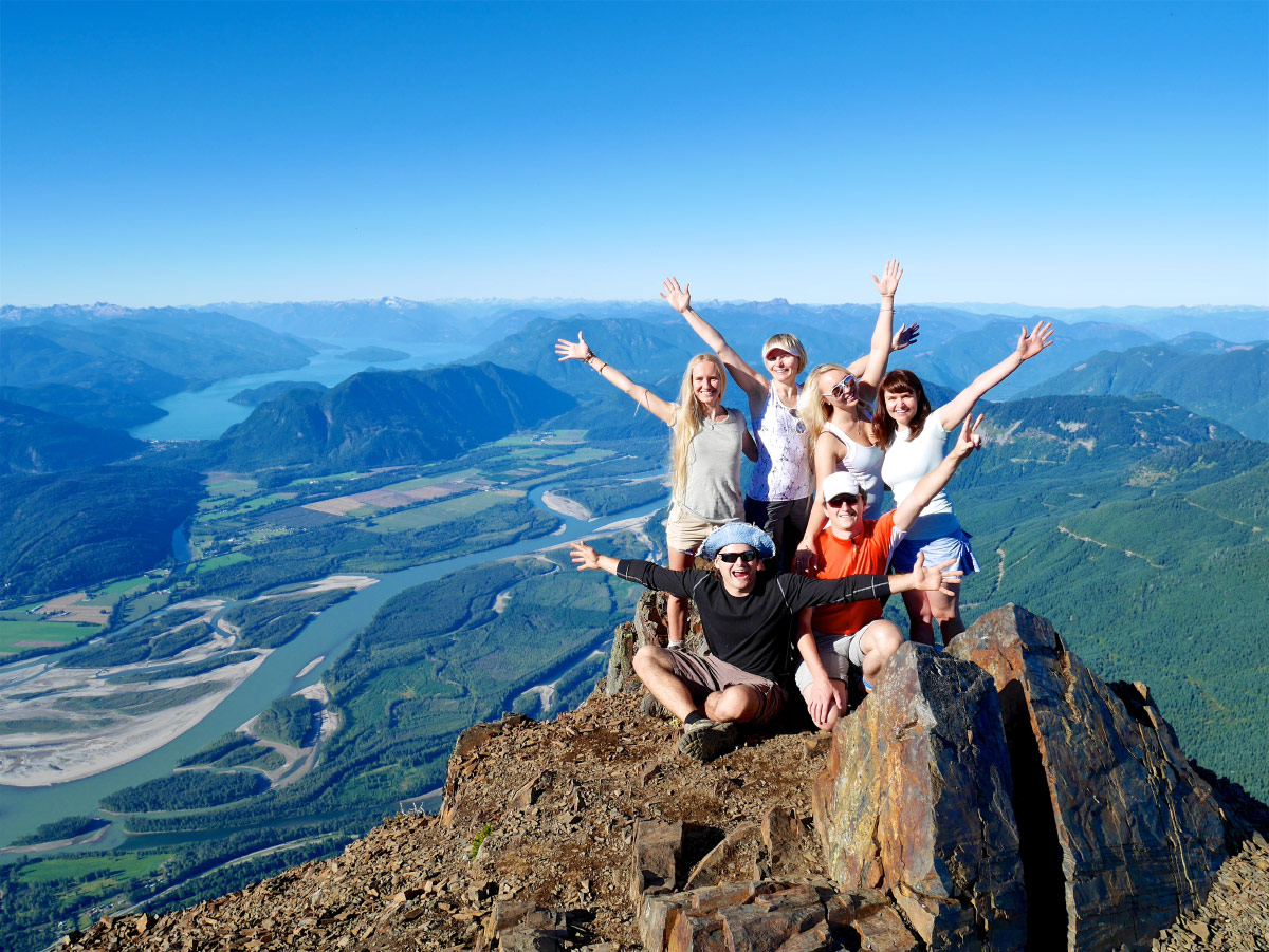 Group of hikers celebrating with raised arms on a mountain summit above a river valley.