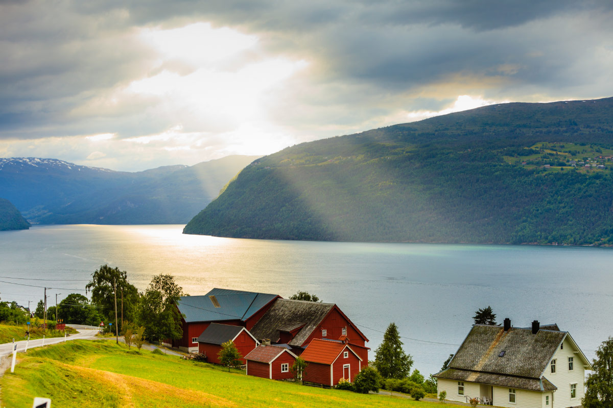 Red farmhouses on a green hillside above a Norwegian fjord with sunbeams through cloud