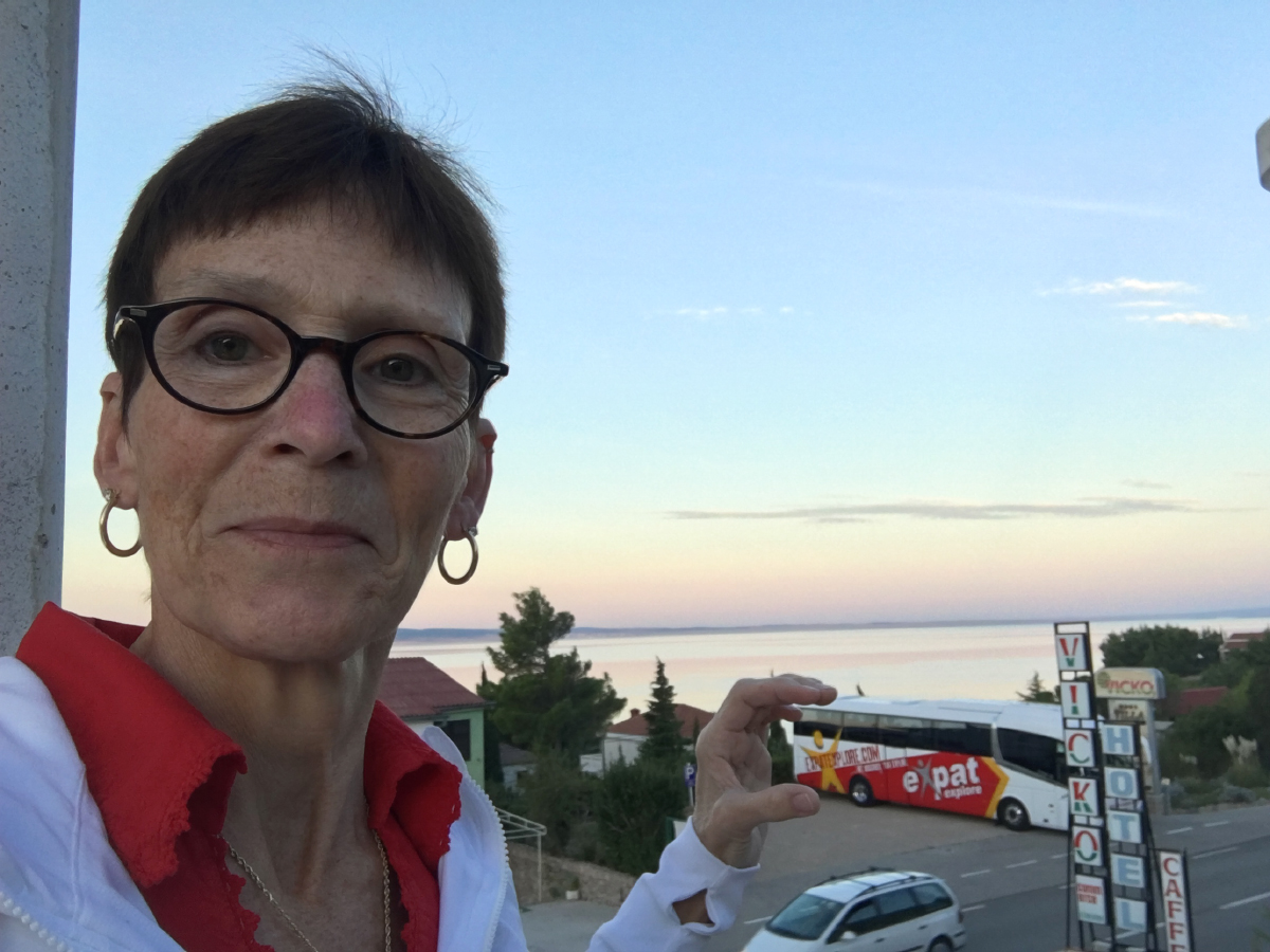Woman in glasses and a red collar smiling beside a coach parked by the sea at sunset