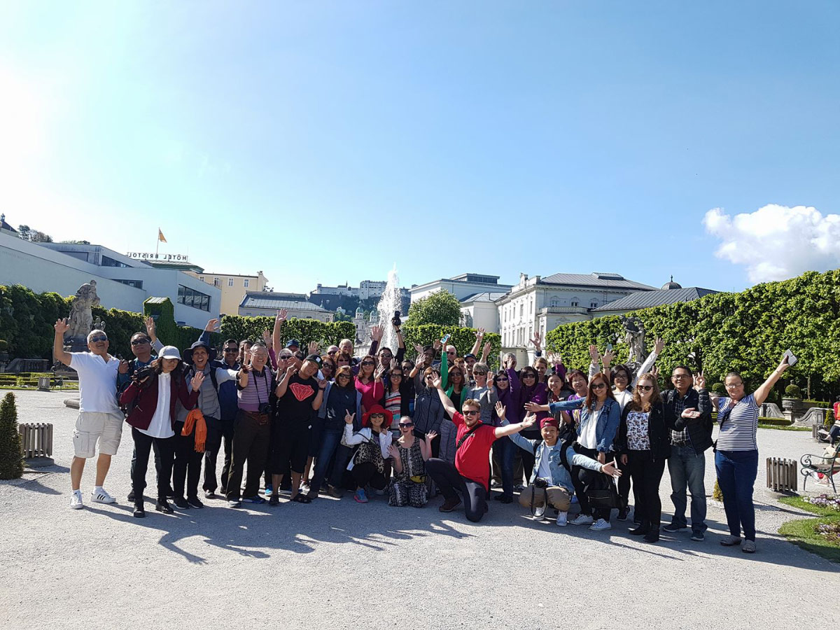 Large group waving in a formal garden with hedges and white buildings behind