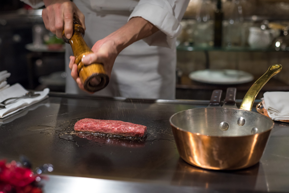 Chef seasoning a wagyu steak on a teppanyaki grill beside a copper pan
