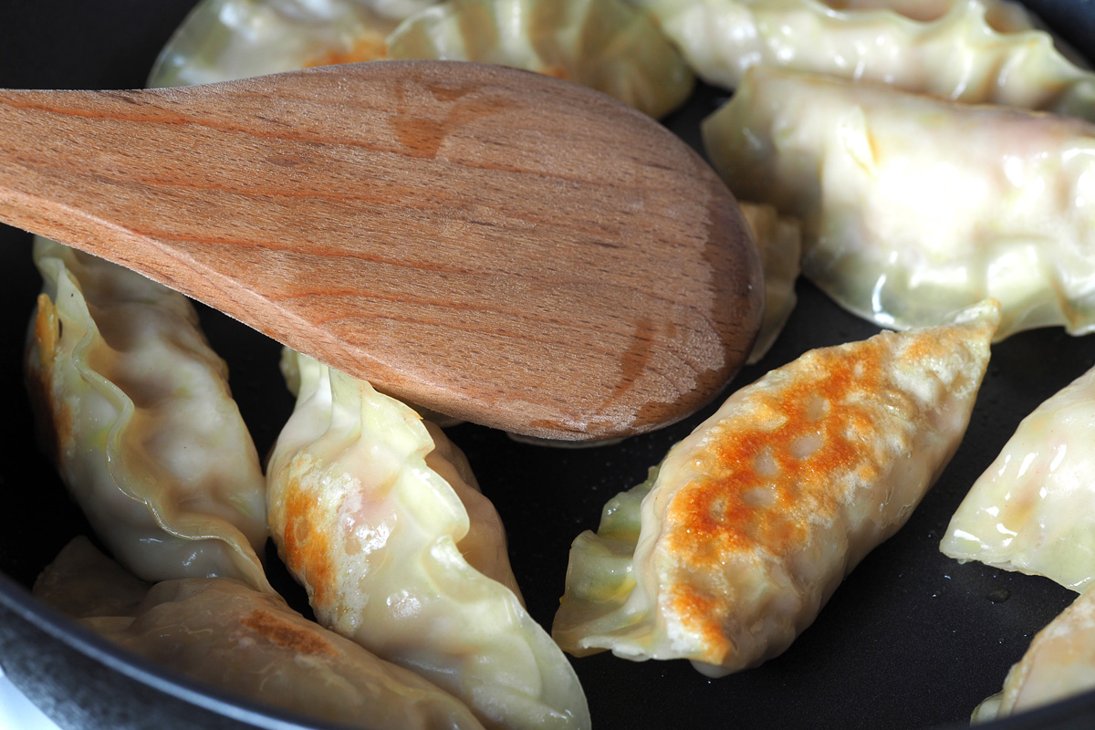 Gyoza dumplings browning in a frying pan with a wooden spatula
