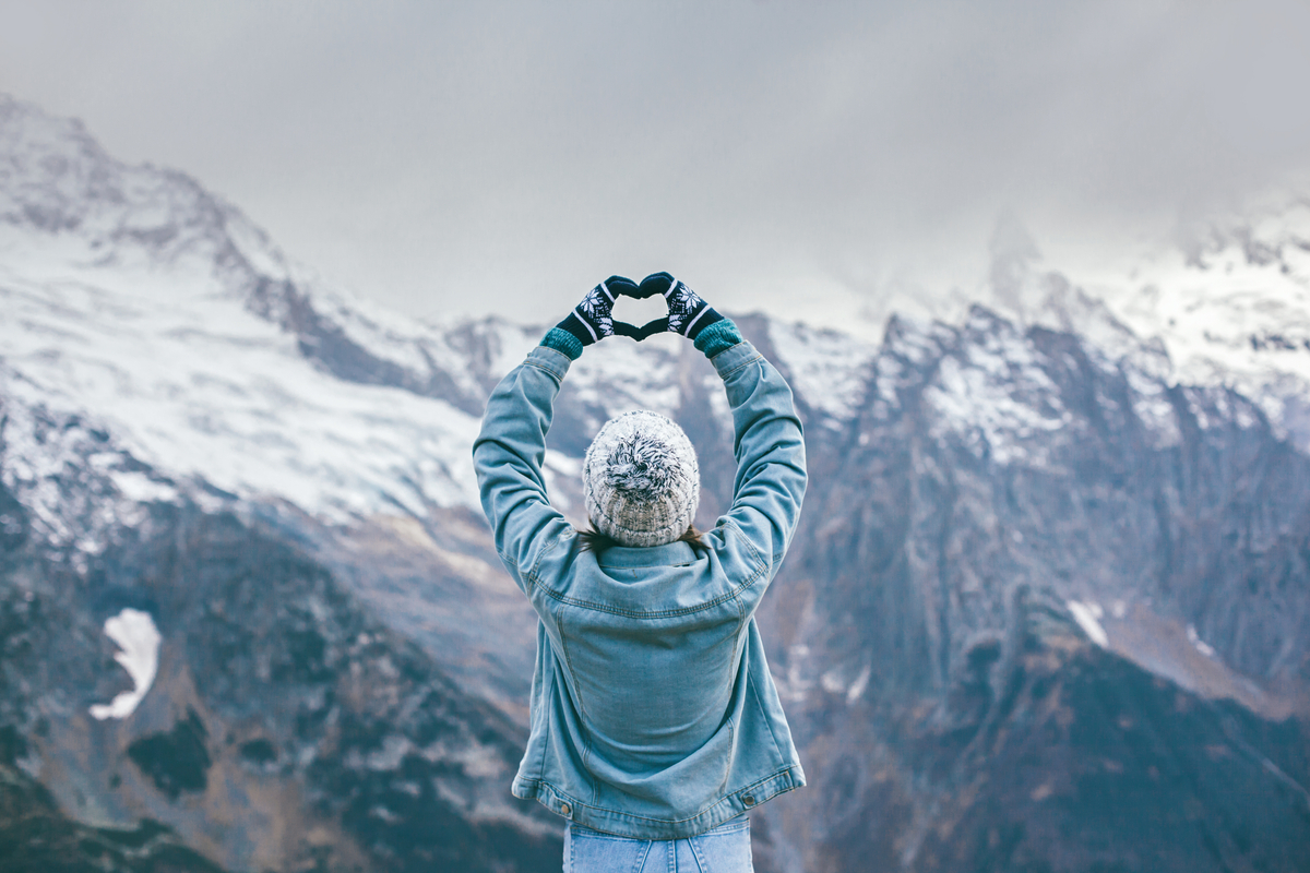 Woman in a knitted hat making a heart with her hands towards snowy mountains