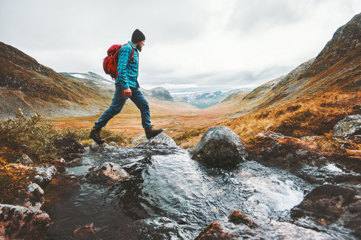 Hiker with a red backpack stepping across a stream in an autumn mountain valley