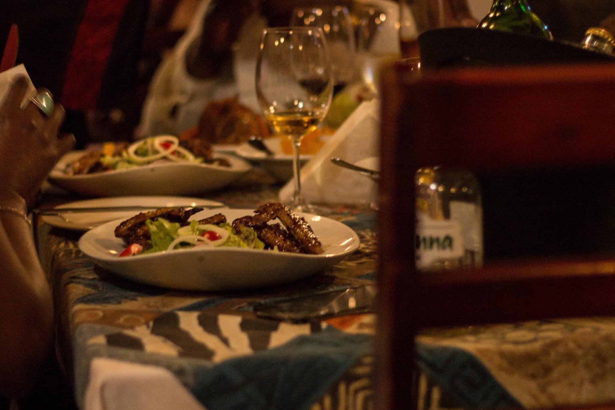 Plates of grilled meat and salad on a patterned tablecloth beside wine glasses